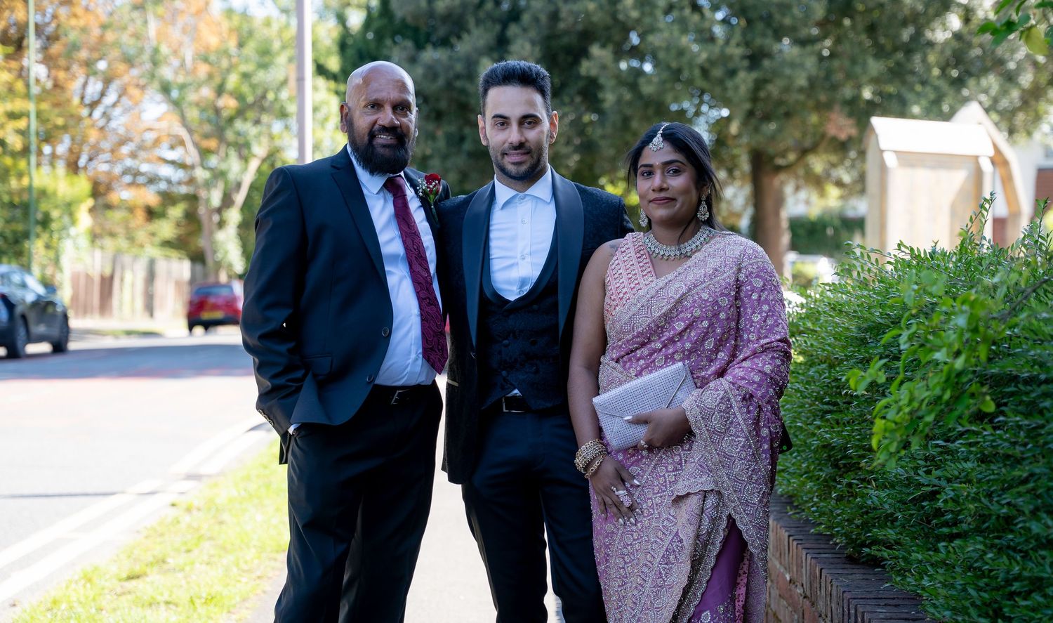 Two men in suits and a woman in pink saree walk together along a tree-lined street at a wedding.