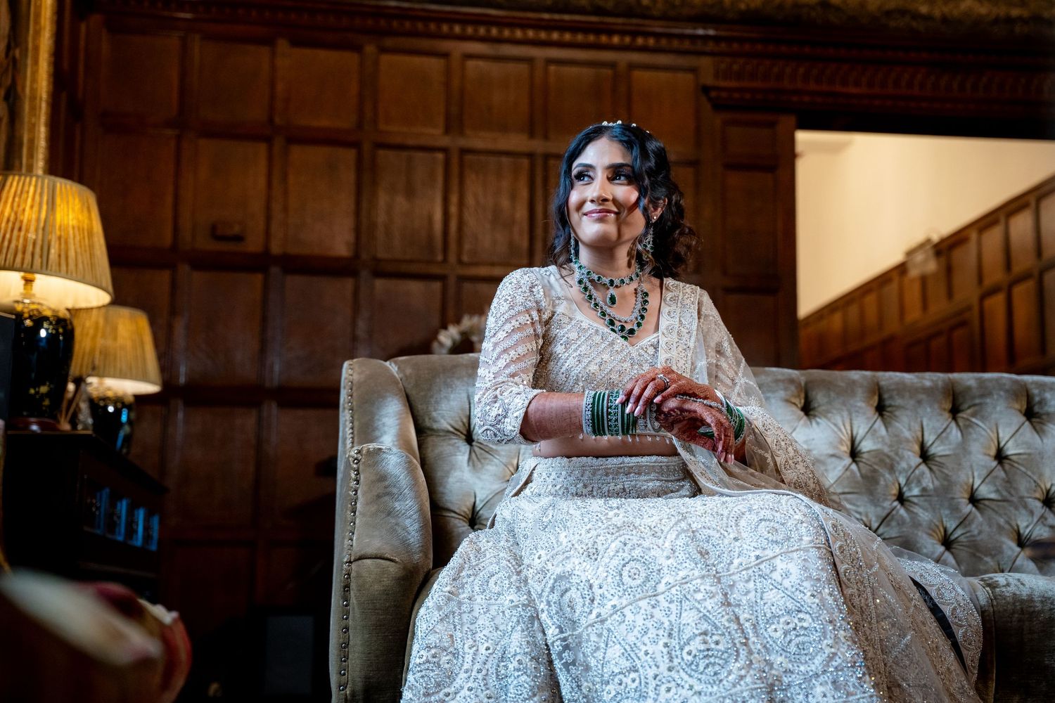 Bride in ivory embroidered lehenga sits gracefully on a tufted sofa in a wood-paneled room.