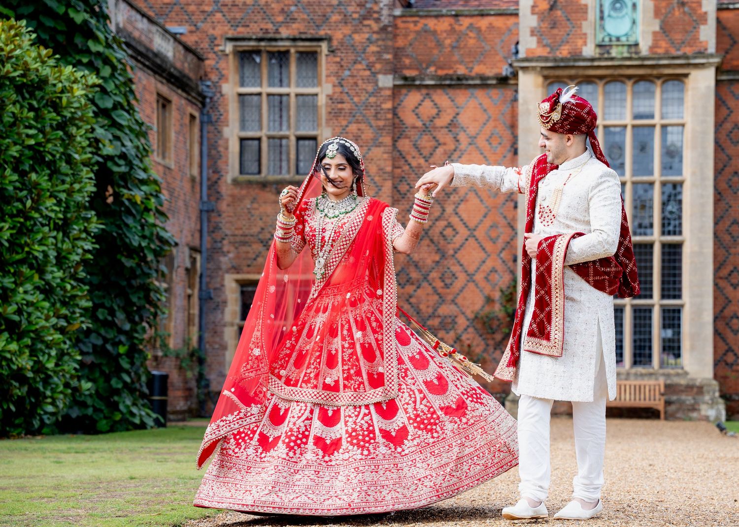 Indian bride in red lehenga and groom in white sherwani dancing outdoors at a traditional wedding ceremony.