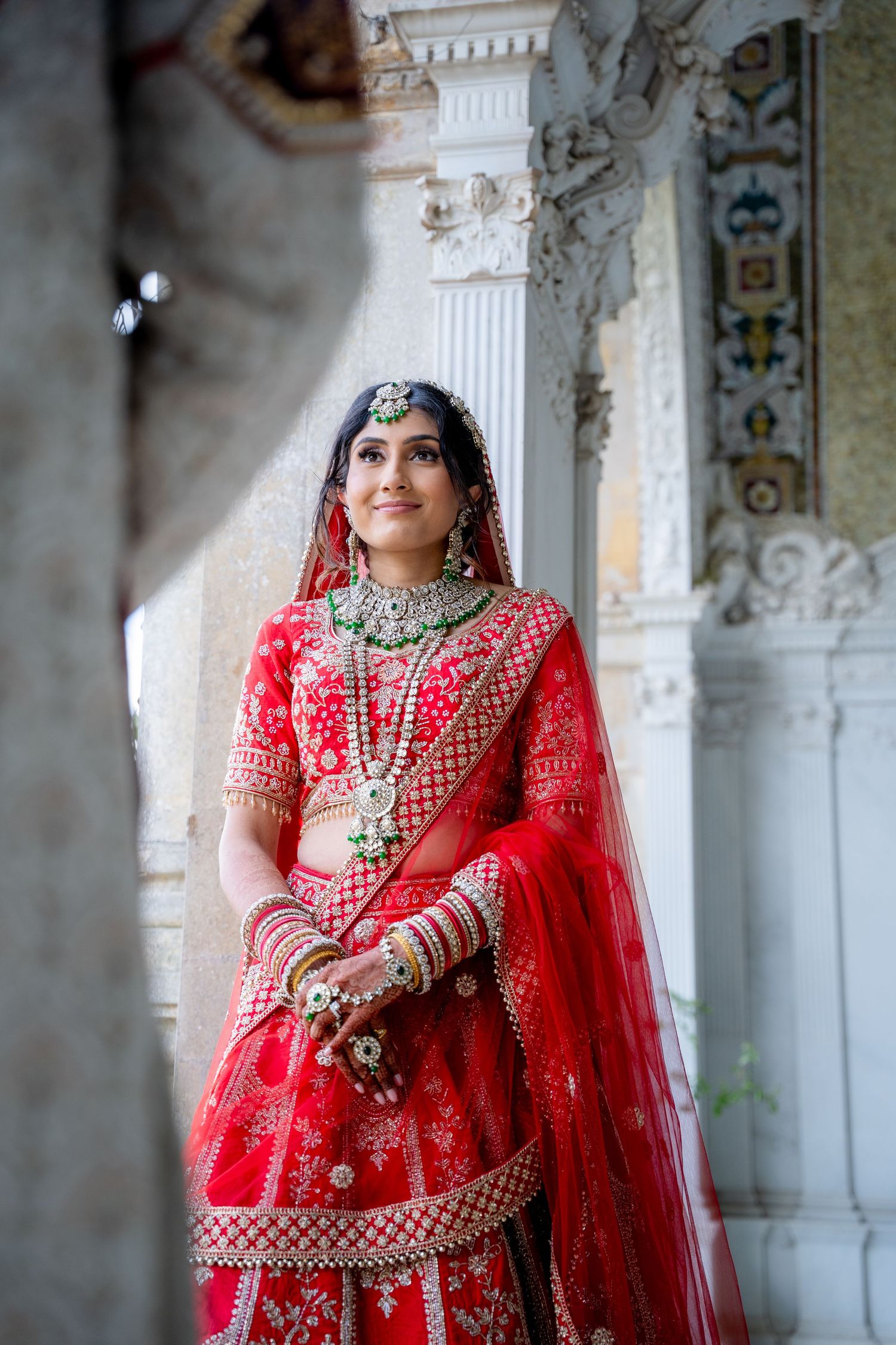 Indian bride in red lehenga with gold embroidery and traditional jewelry poses in ornate white palace.