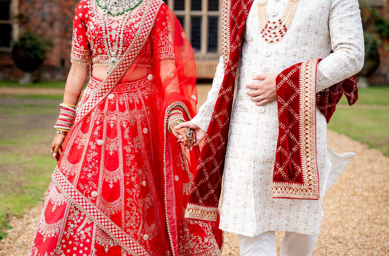 Close-up of Indian bride and groom holding hands in intricate red lehenga and white sherwani wedding attire.