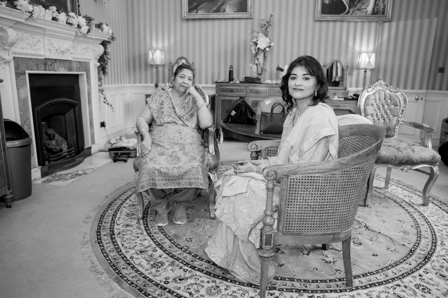 Black and white photo of two women in sarees seated in an elegant drawing room.