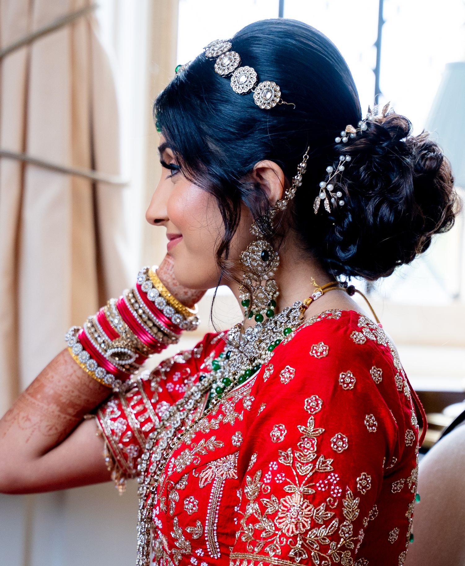 Indian bride smiles with ornate silver hair accessories and bangles in red bridal lehenga.