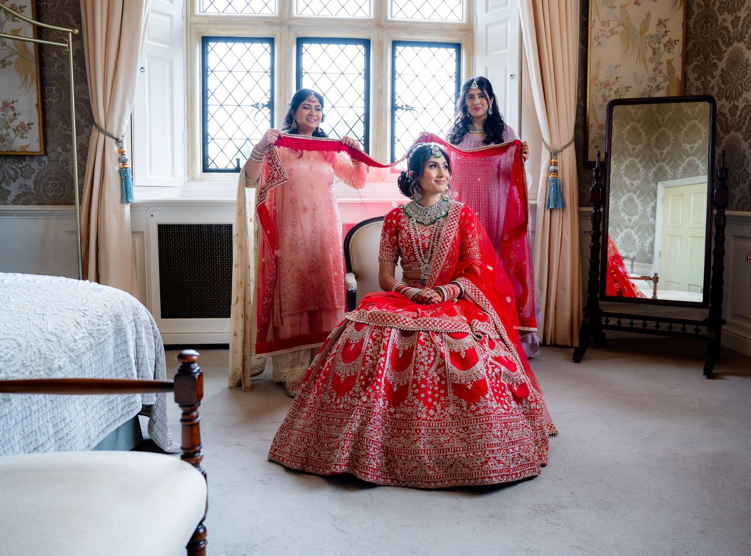 Indian bride in red lehenga seated while two bridesmaids hold her dupatta in elegant bedroom.