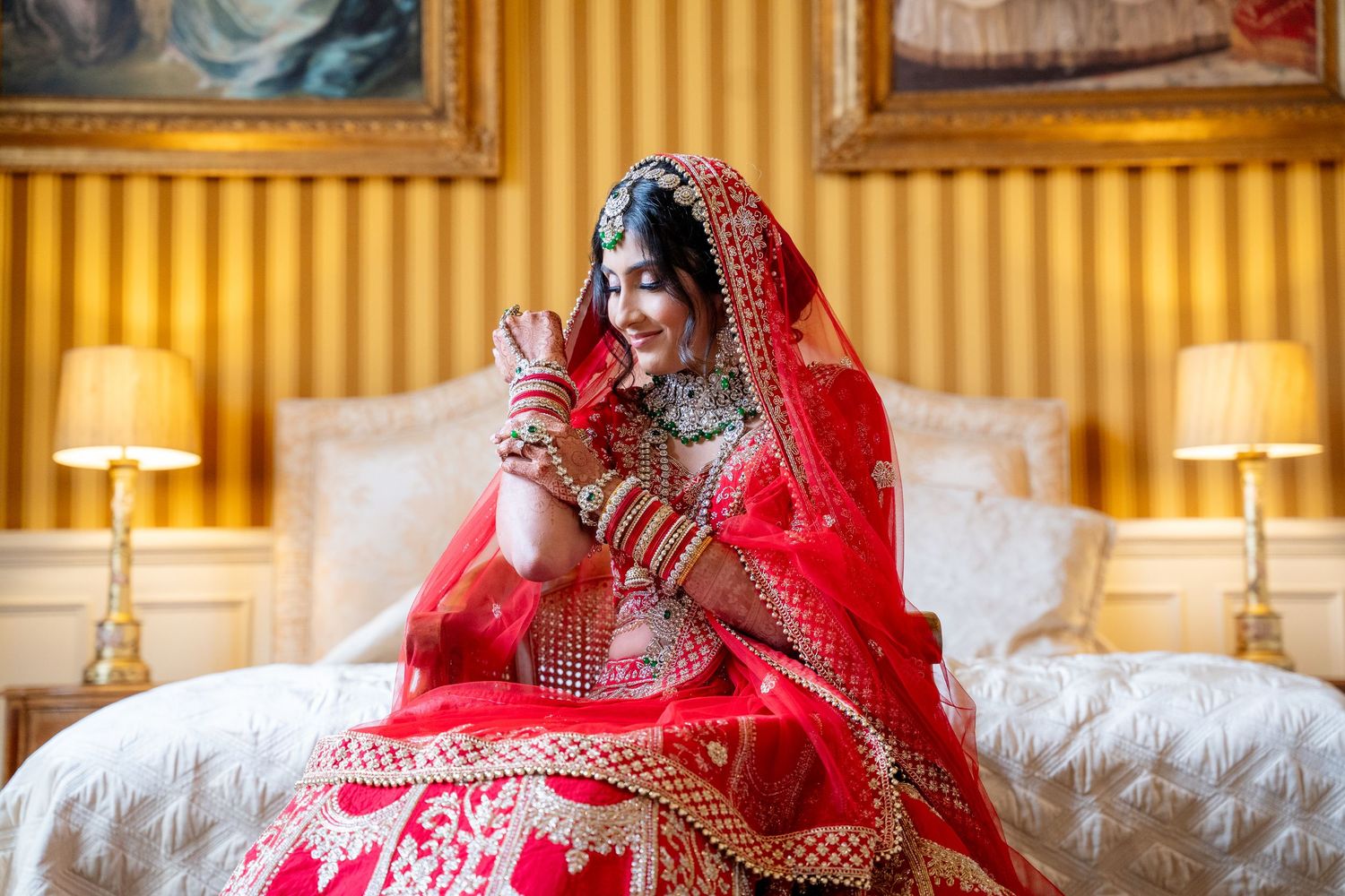 Indian bride in red embroidered lehenga sitting on bed admiring her bridal jewelry.