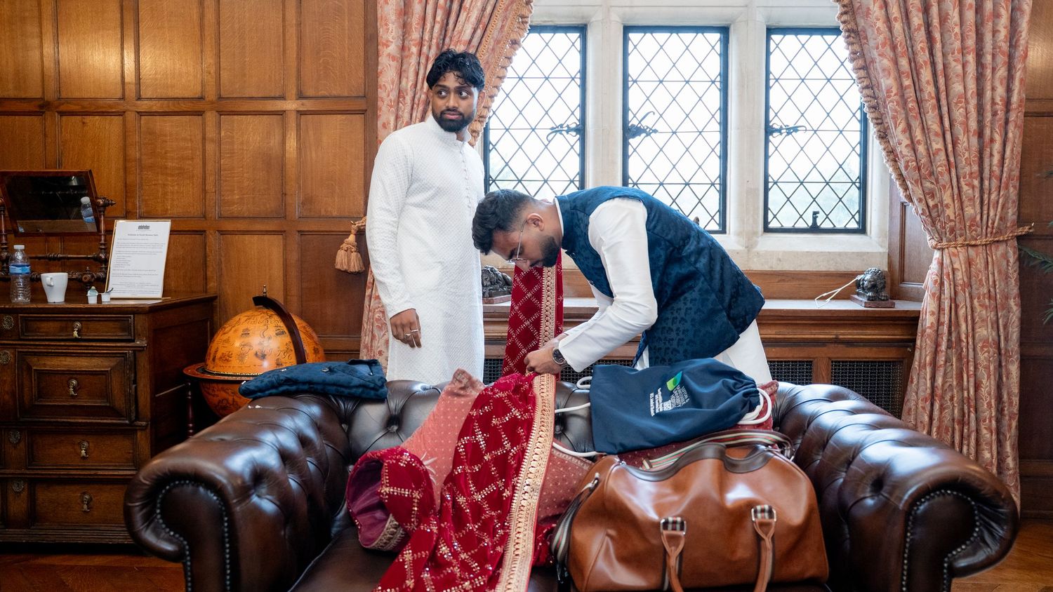Indian groomsmen in traditional attire arranging red wedding outfit on a leather Chesterfield sofa.