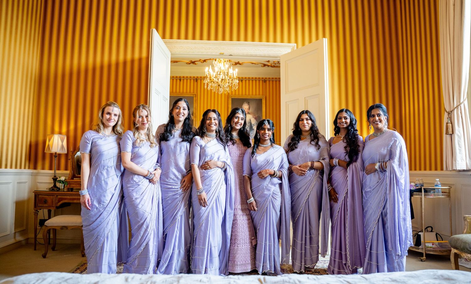 Nine bridesmaids in matching lavender sarees posing together in golden-walled elegant wedding room.
