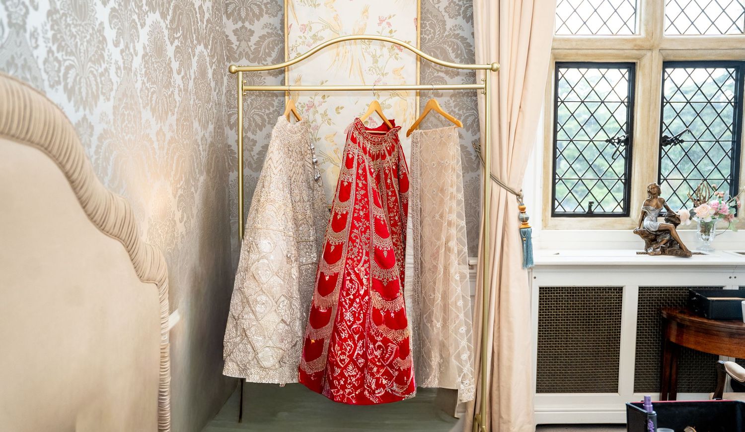 Wedding outfits including white embroidered lehenga and red bridal dress hanging on a gold rack.