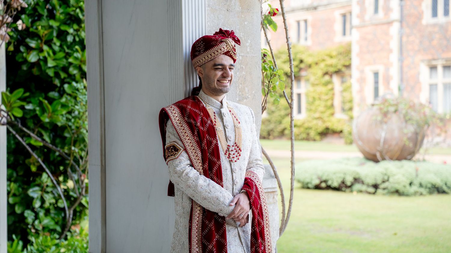 Groom in traditional Indian wedding attire with red sherwani and turban posing by a white pillar.