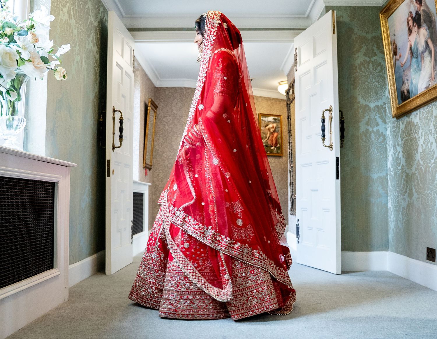 Indian bride in stunning red embroidered lehenga with veil standing in elegant hallway.