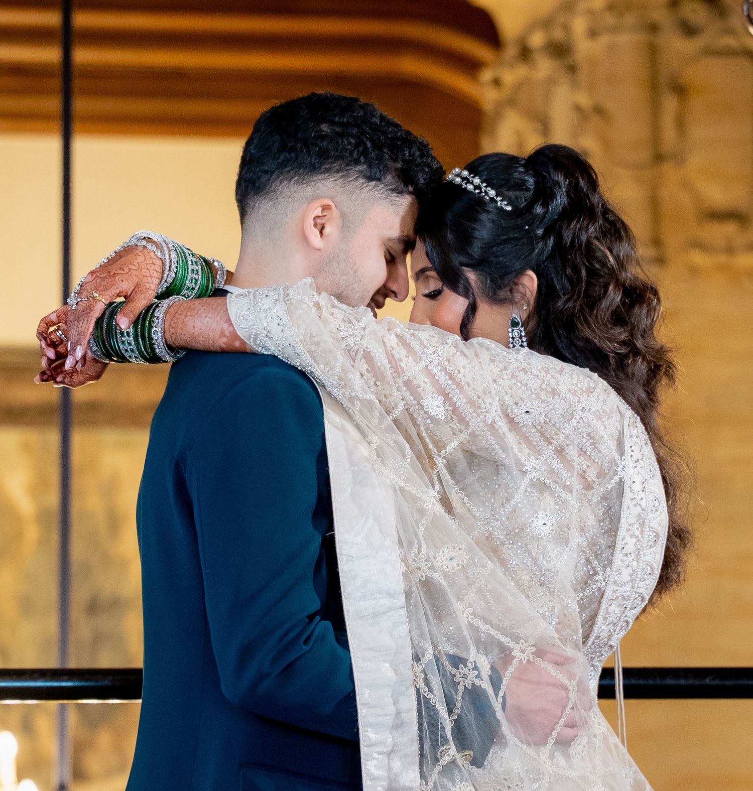 South Asian bride in white embroidered lehenga with green bangles embraces groom in navy suit at wedding.