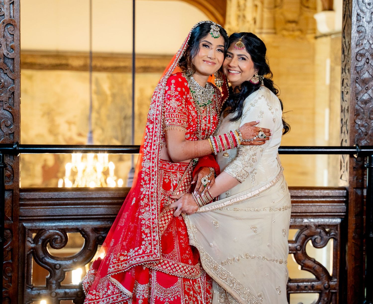 Indian bride in red lehenga embraces woman in cream saree at decorated wedding venue.
