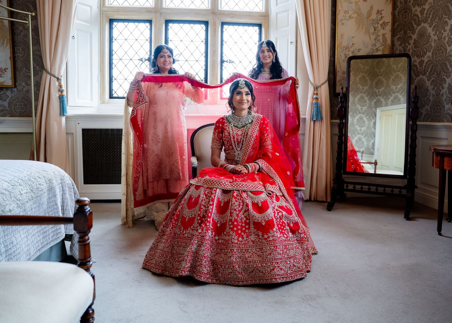 Indian bride in red lehenga seated while two bridesmaids hold her dupatta in an elegant room.
