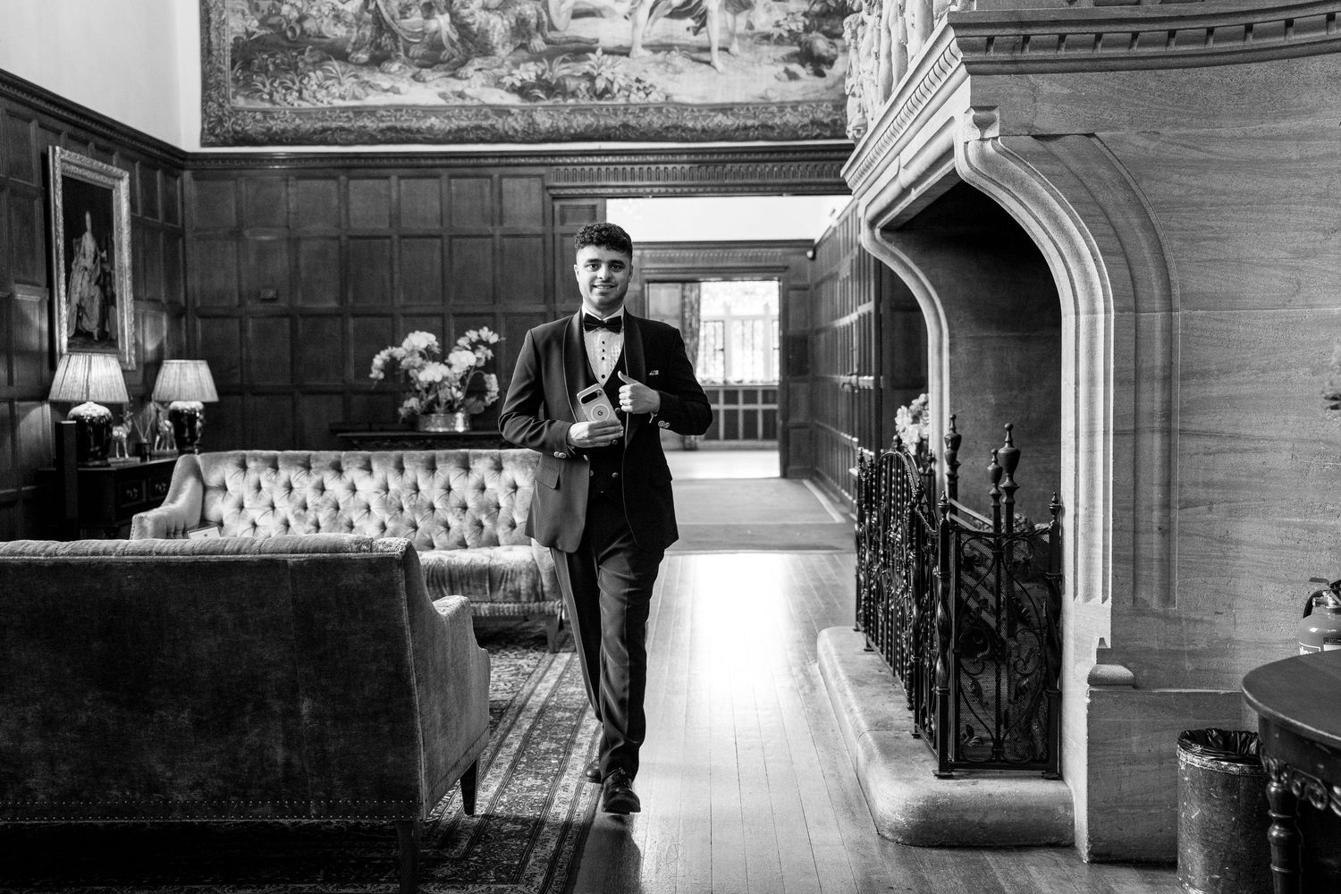 A groom in a black tuxedo and bow tie walks confidently through an elegant wood-paneled hall.
