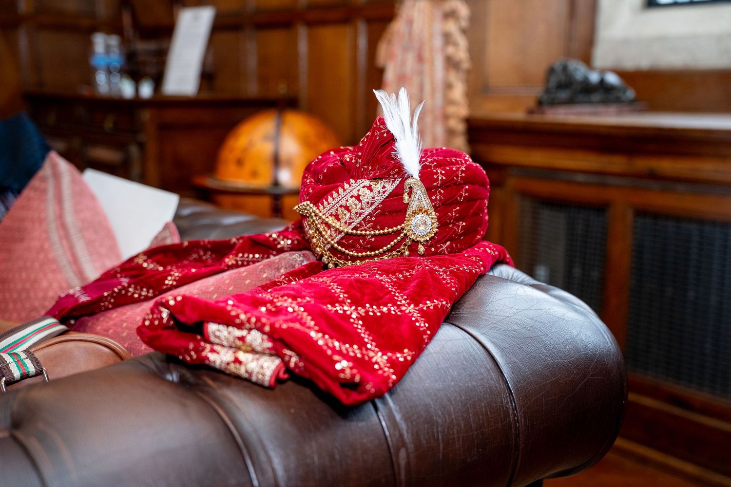 Red embroidered Indian groom turban with white feather plume resting on brown leather sofa.