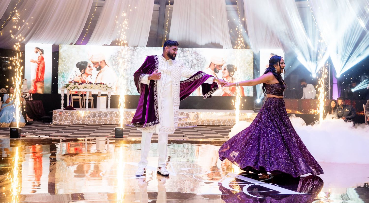 Couple performs traditional dance in purple attire at an elegant wedding reception with decorative lighting and stage setup.