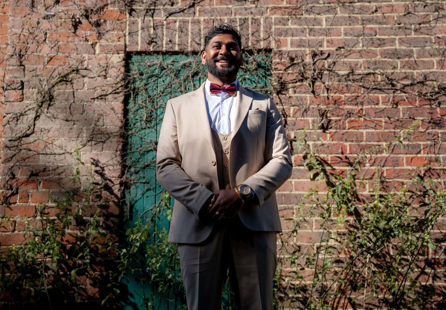 Groom in tan suit and red bow tie posing confidently against a rustic brick wall with ivy.