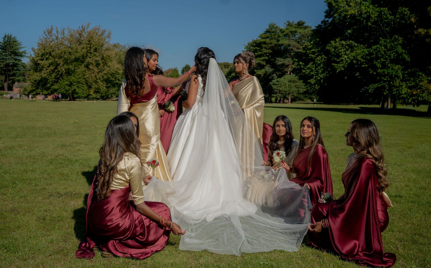 Bridesmaids in burgundy and gold dresses arrange brides veil on sunny green lawn at wedding.