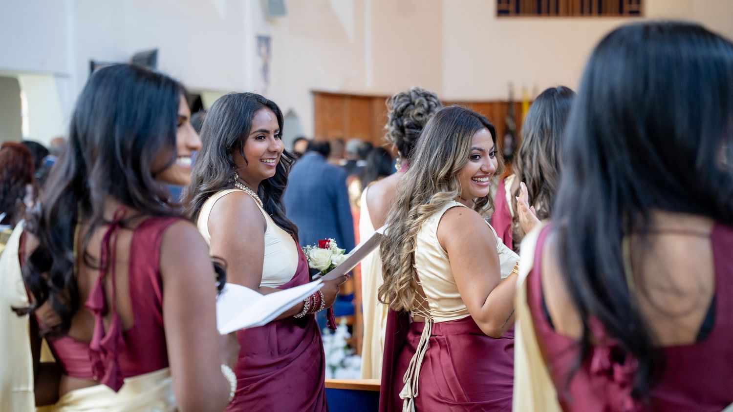 Bridesmaids in burgundy and gold saris laugh together inside wedding venue before ceremony.