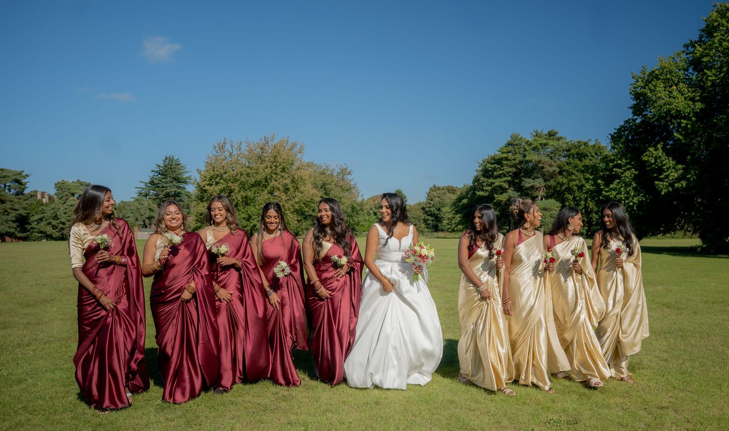 Bride in white gown with bridesmaids in red and gold satin dresses posing outdoors on green lawn under blue sky.