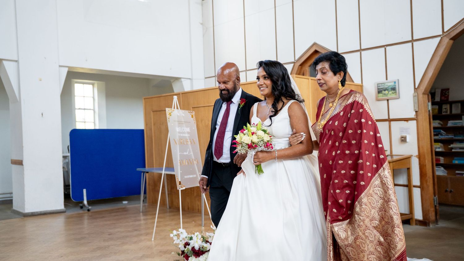 Bride in white dress walks with escort and woman in red saree through a bright modern church ceremony.