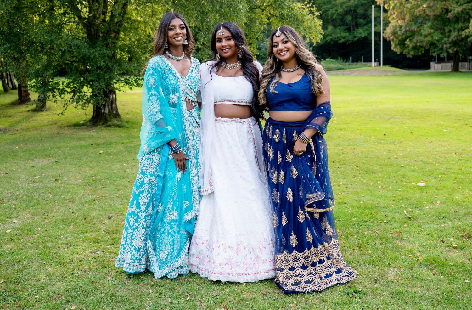 Three women in vibrant Indian lehenga outfits posing together in a lush green park setting.