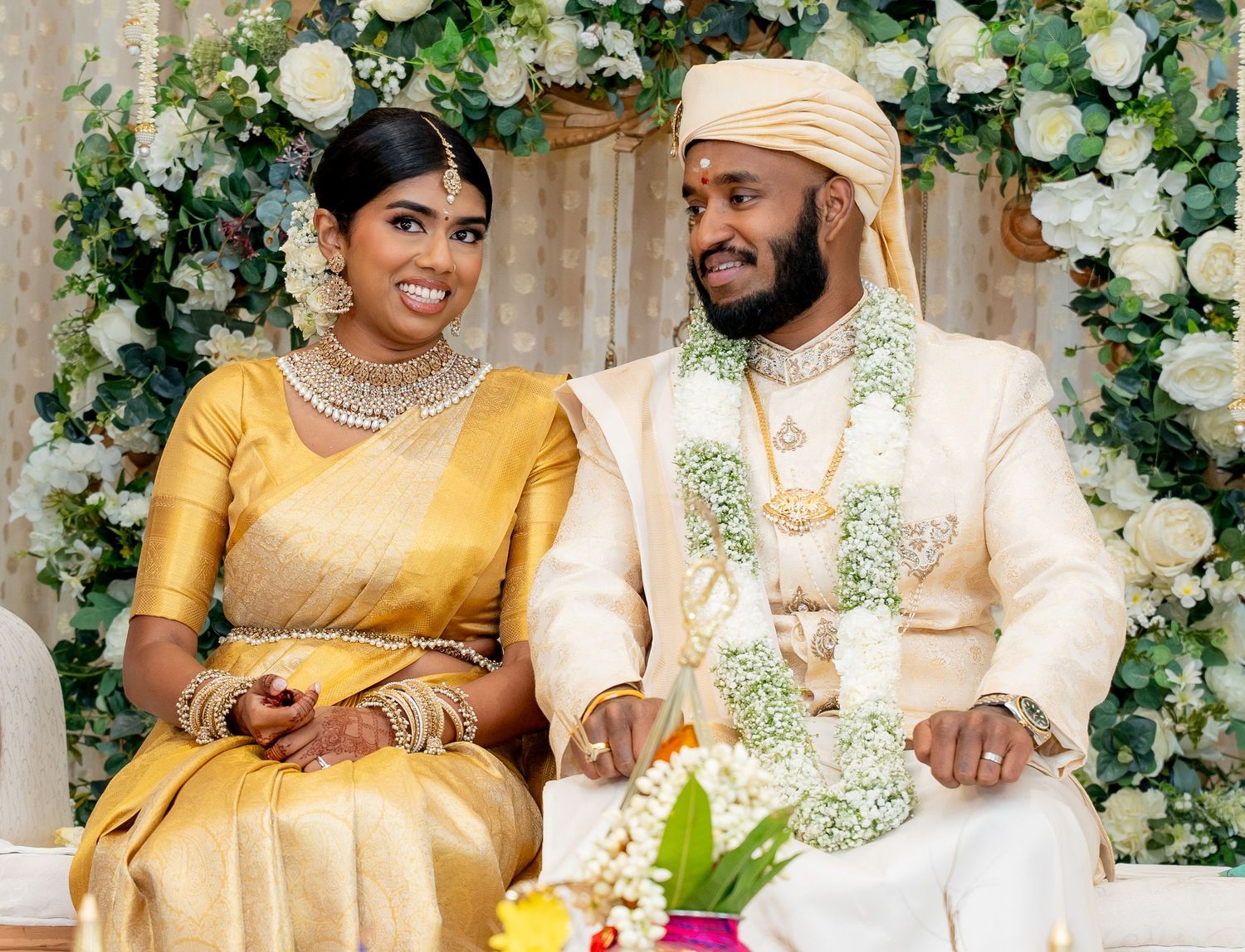 Couple in traditional South Asian wedding attire, bride in gold saree with jewelry, groom in white sherwani, seated at ceremony.