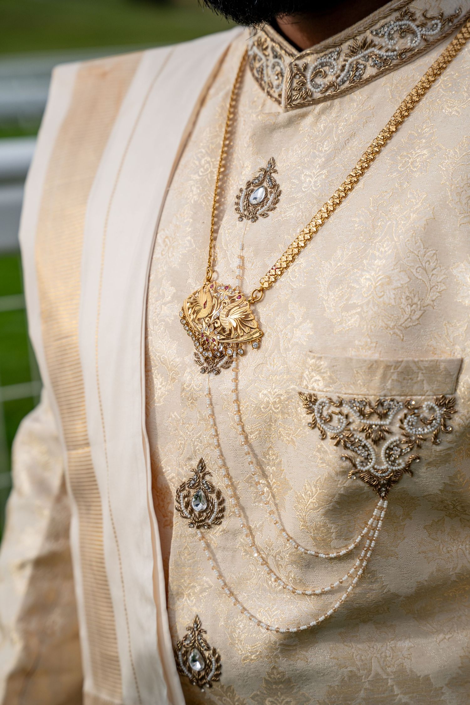 Man wearing cream brocade sherwani with gold embroidery and layered gold necklace at Indian wedding ceremony.
