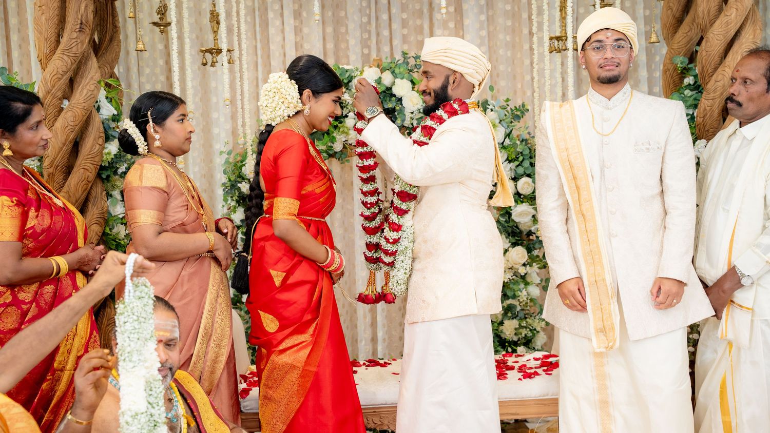 Groom places a floral garland on bride in red saree during a colorful South Indian wedding ritual.