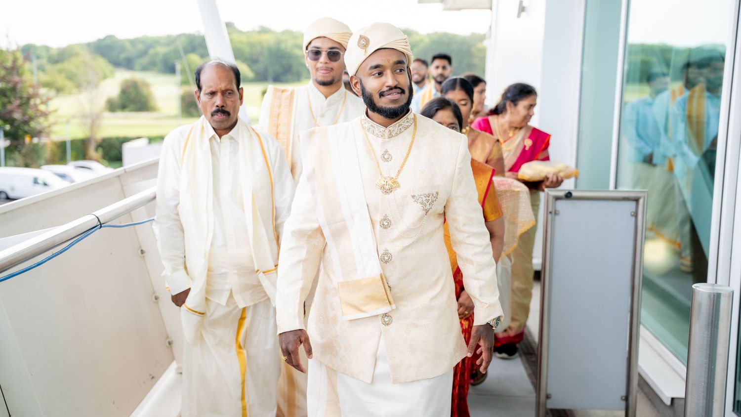 Two grooms in elegant white and gold Indian wedding outfits lead a wedding procession through a modern venue.