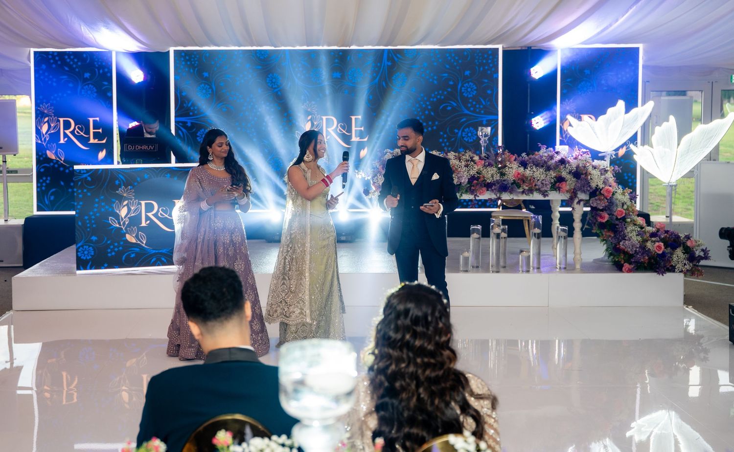 Elegant wedding reception with couple on stage, floral decor, blue lighting, and monogram backdrop at a formal venue.