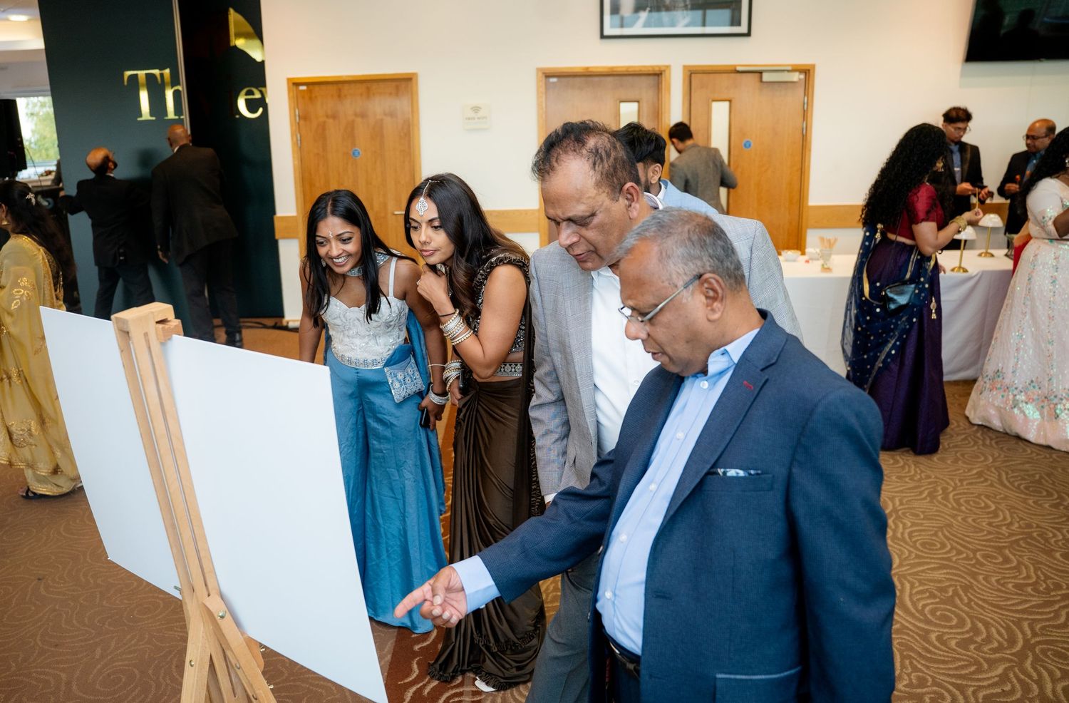 Guests in formal attire examine a presentation board on an easel at an elegant indoor social event or gala.