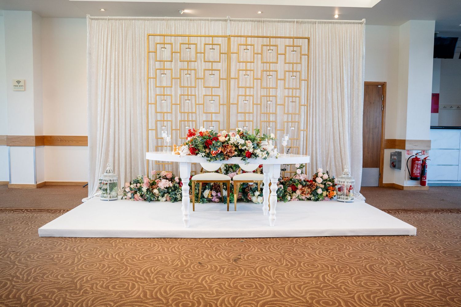 Elegant wedding sweetheart table with floral arrangements on a white stage with decorative backdrop.