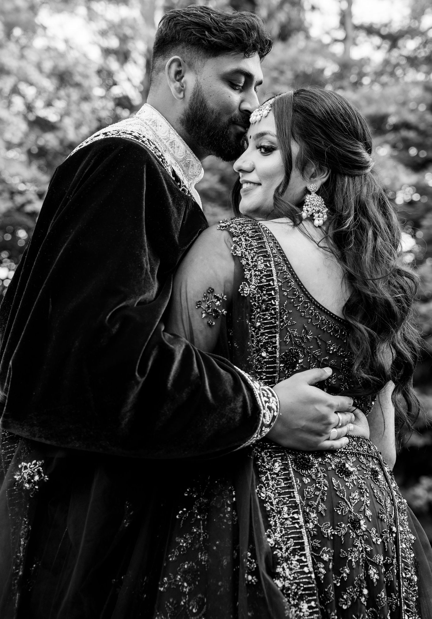 Black and white portrait of South Asian bride and groom in embellished traditional attire embracing outdoors.