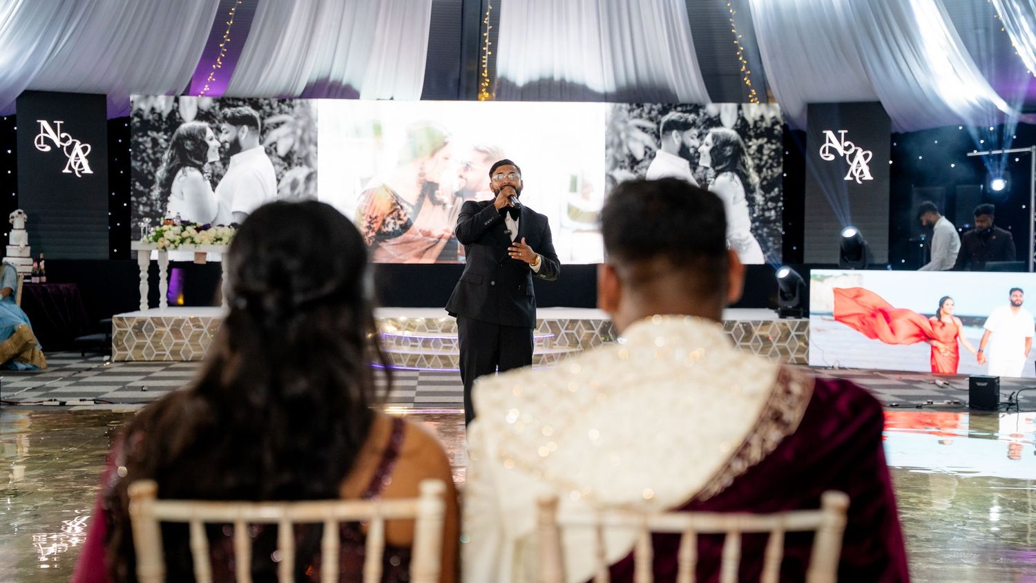 A speaker addresses a couple at an elegant Indian wedding reception with floral decor and a lit backdrop.