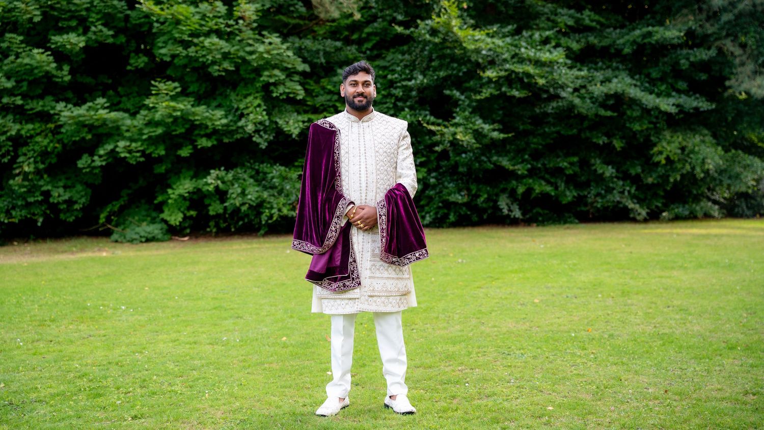 Man in traditional South Asian sherwani with purple dupatta standing on green lawn with trees.