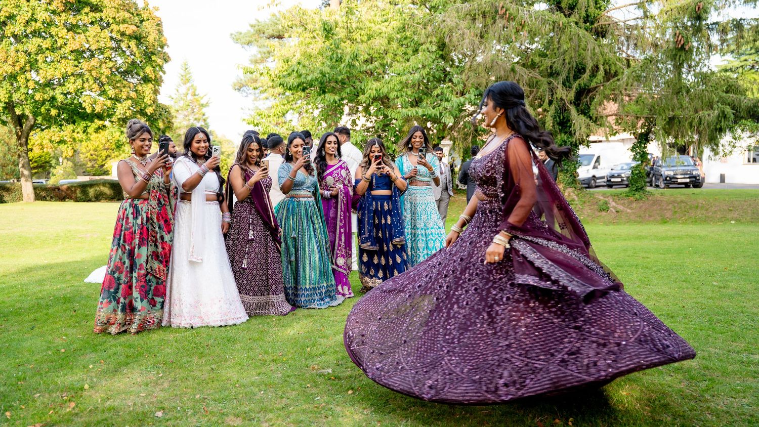 Bride in purple lehenga twirling elegantly on a green lawn while wedding guests cheer and watch in colorful attire.