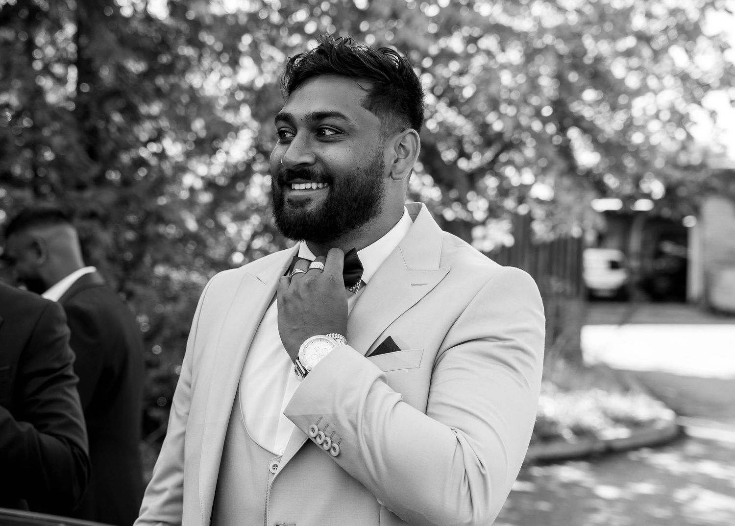 Bearded man in white suit smiles joyfully outdoors at a wedding celebration in black and white.