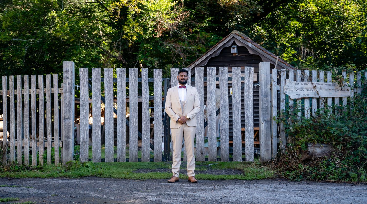 Groom in ivory suit stands before a rustic white picket fence in dappled sunlight.