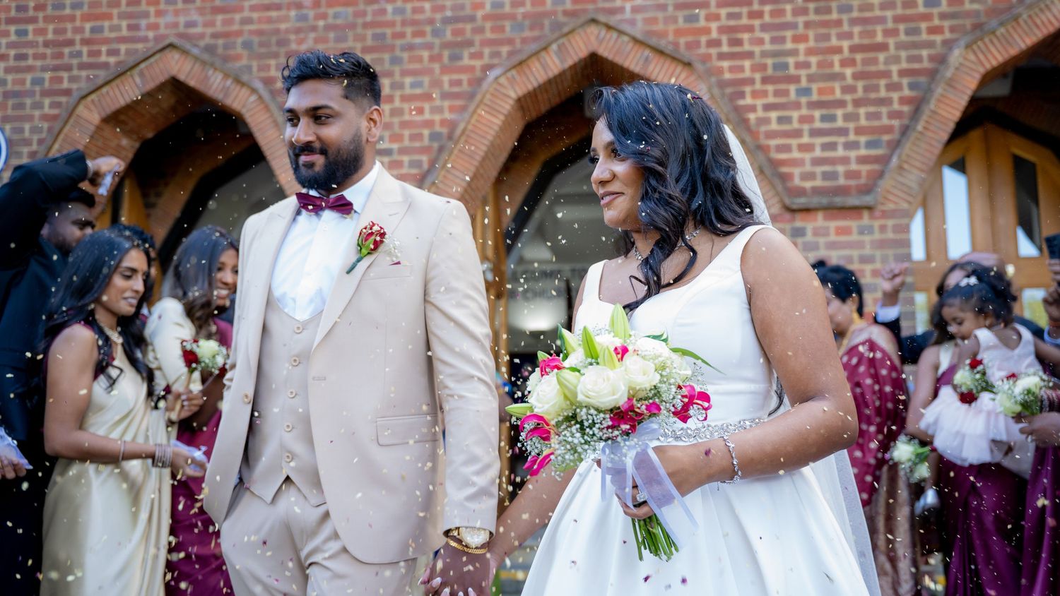 Newlywed couple walks from brick church as guests throw confetti, bridesmaids in burgundy dresses behind.