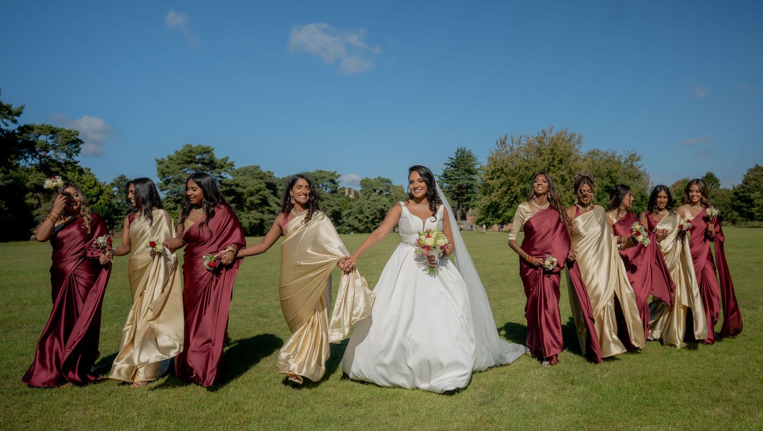 Bride and bridesmaids in burgundy and gold satin dresses posing on green grass under clear sky.