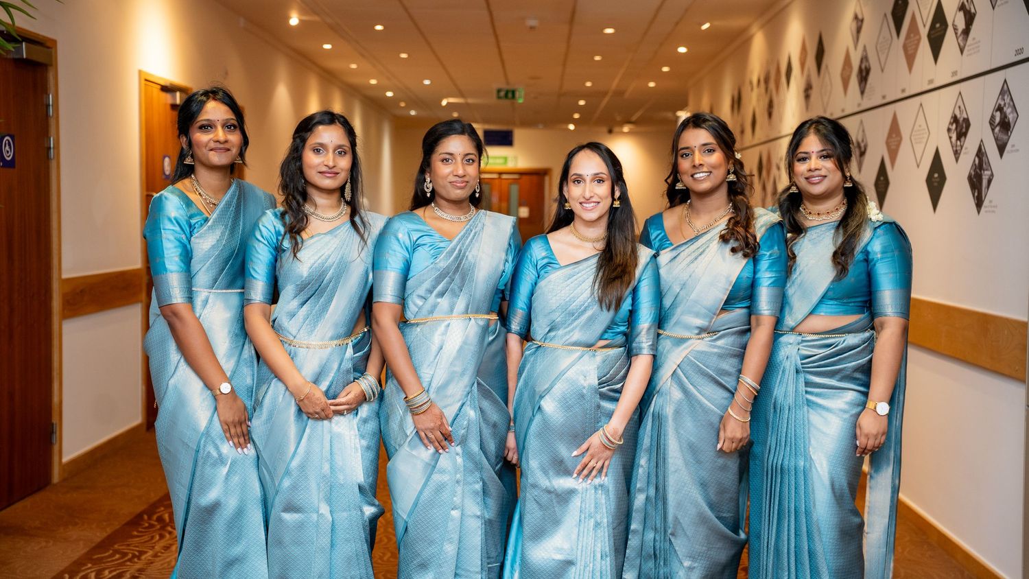 Group of six women wearing coordinating light blue silk sarees posing in a hotel corridor.