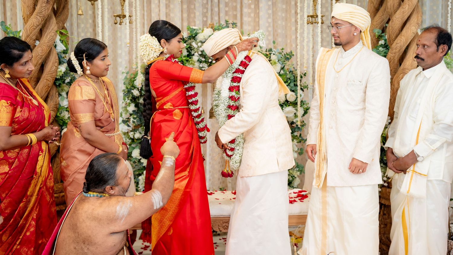 Bride in red silk saree places wedding garland on groom during South Indian Hindu marriage ceremony.