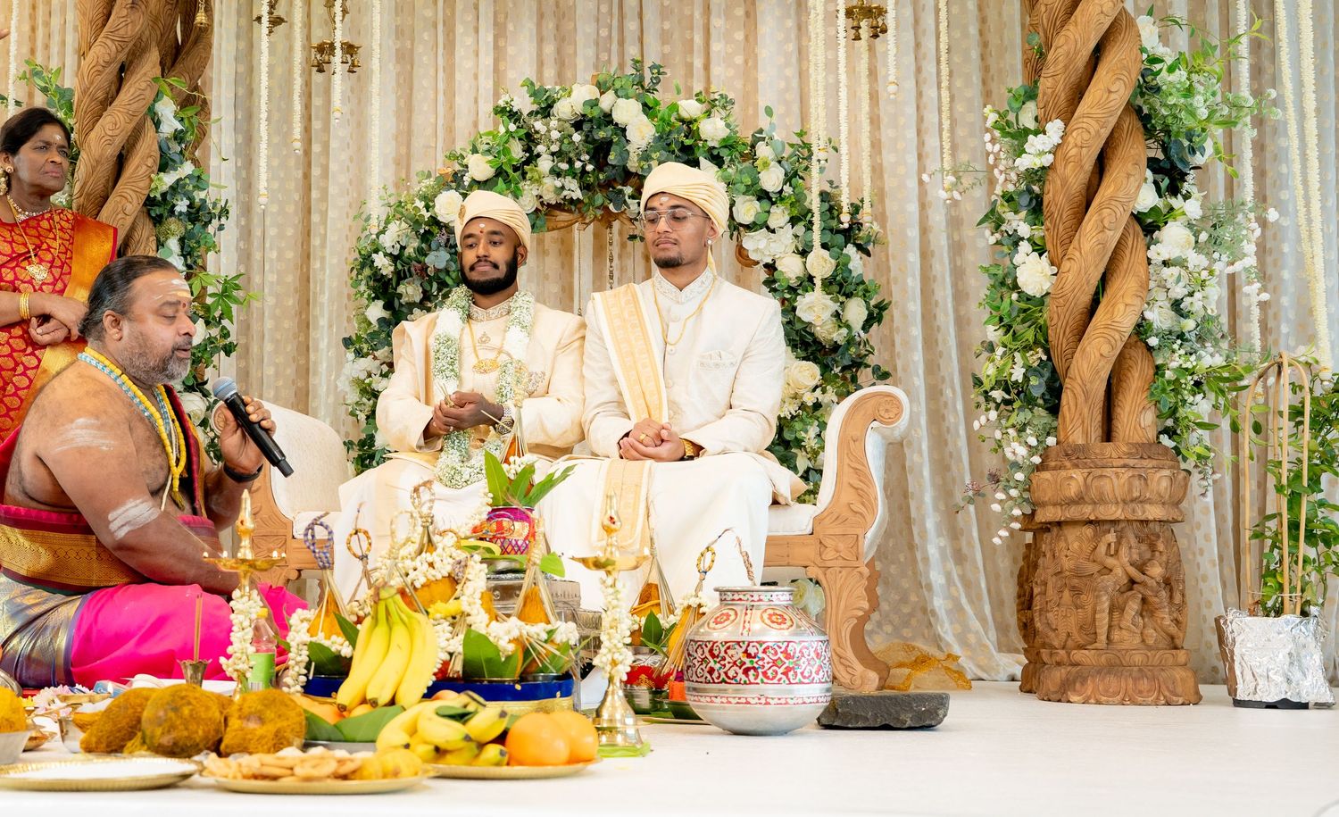 Two grooms in elegant white attire seated at a Hindu wedding ceremony with a speaking priest.