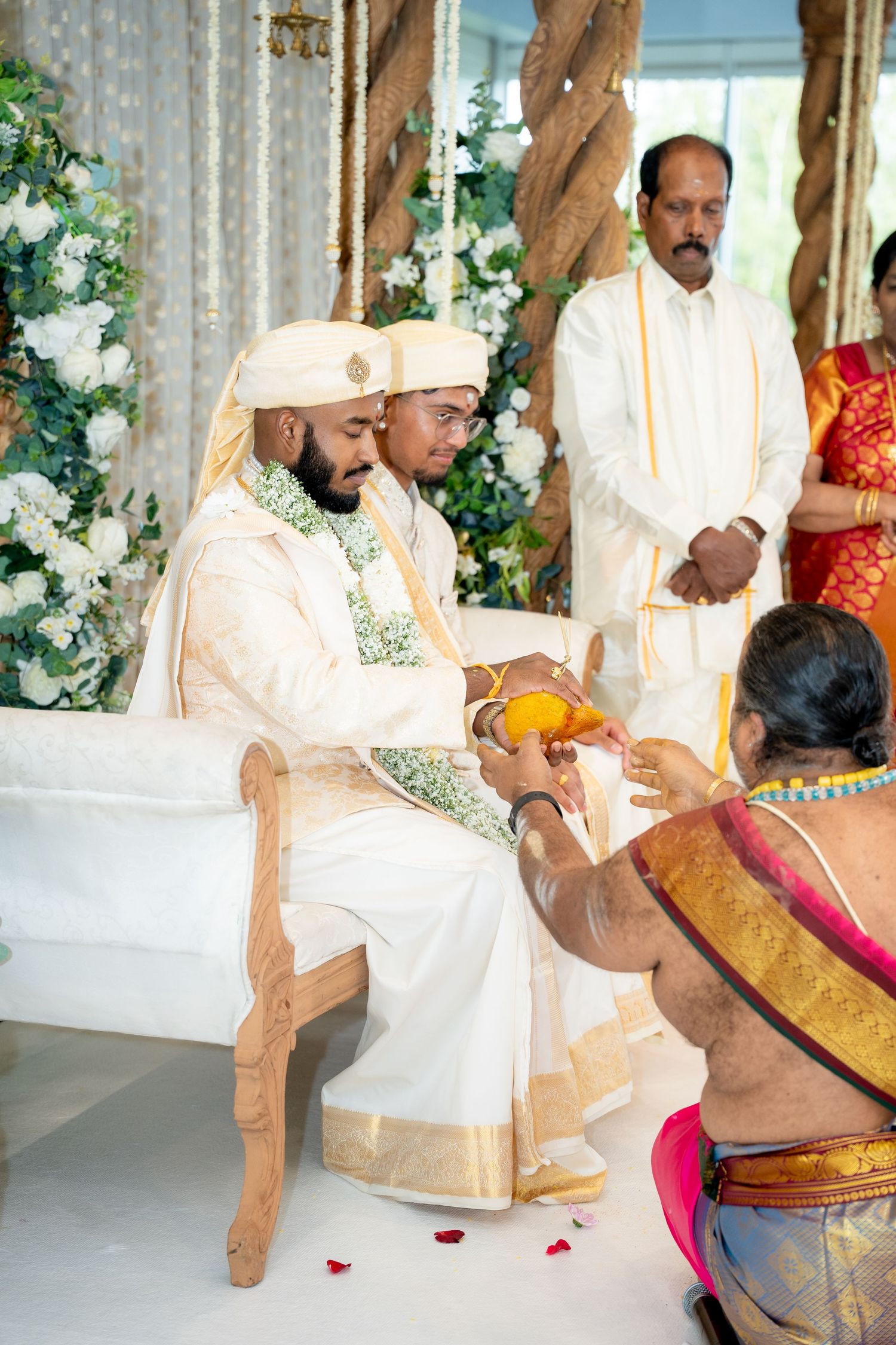 Groom in white traditional attire receiving sacred coconut offering from Hindu priest during South Indian wedding ritual.