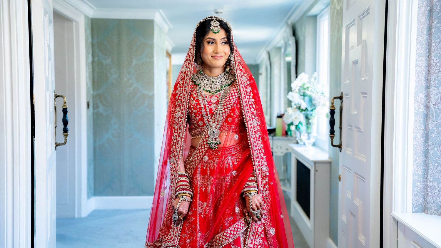 Bride in red lehenga with gold embroidery and traditional jewelry poses in hallway for Indian wedding portrait.