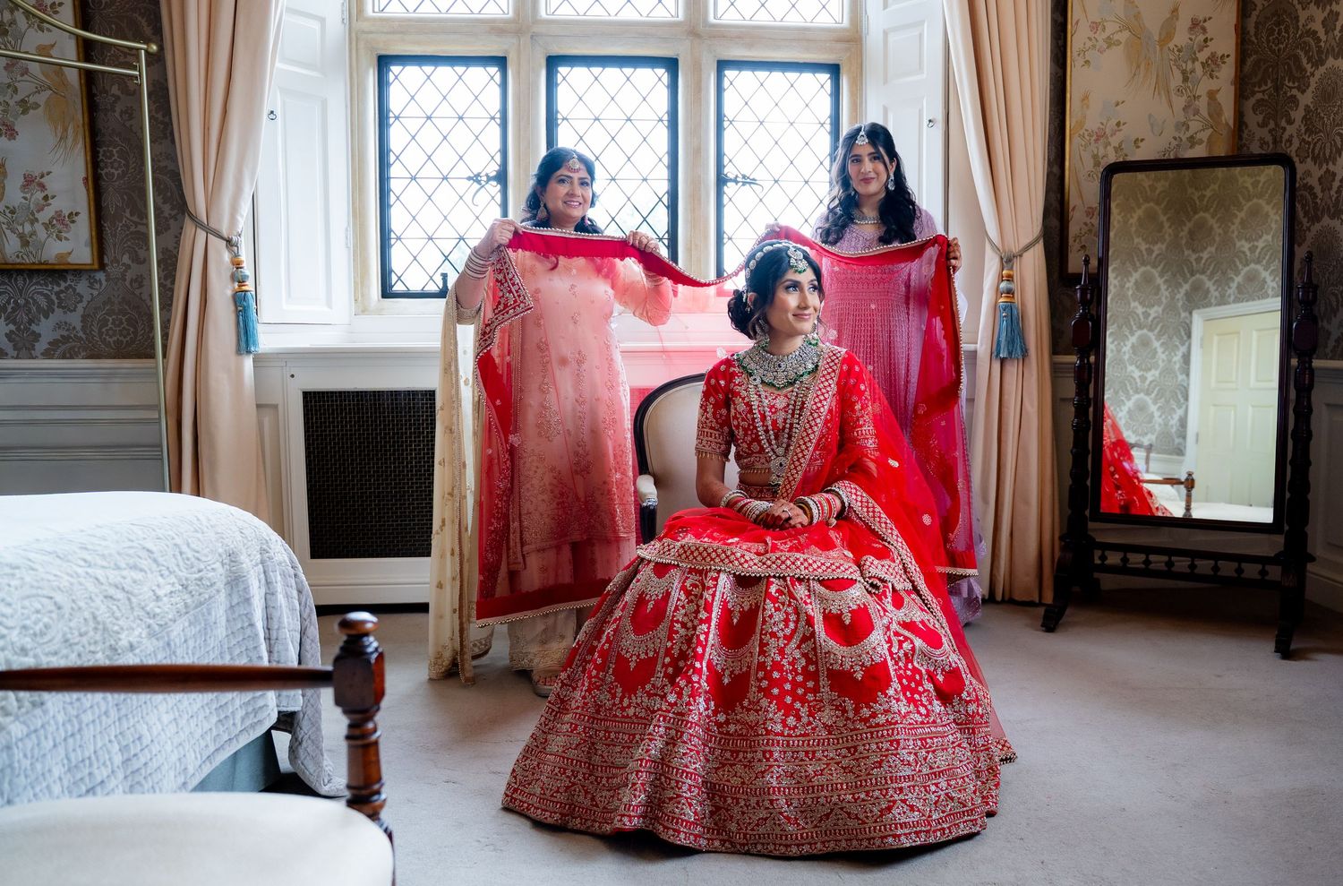Bride in red bridal lehenga sits as two women help arrange her dupatta in a grand room.