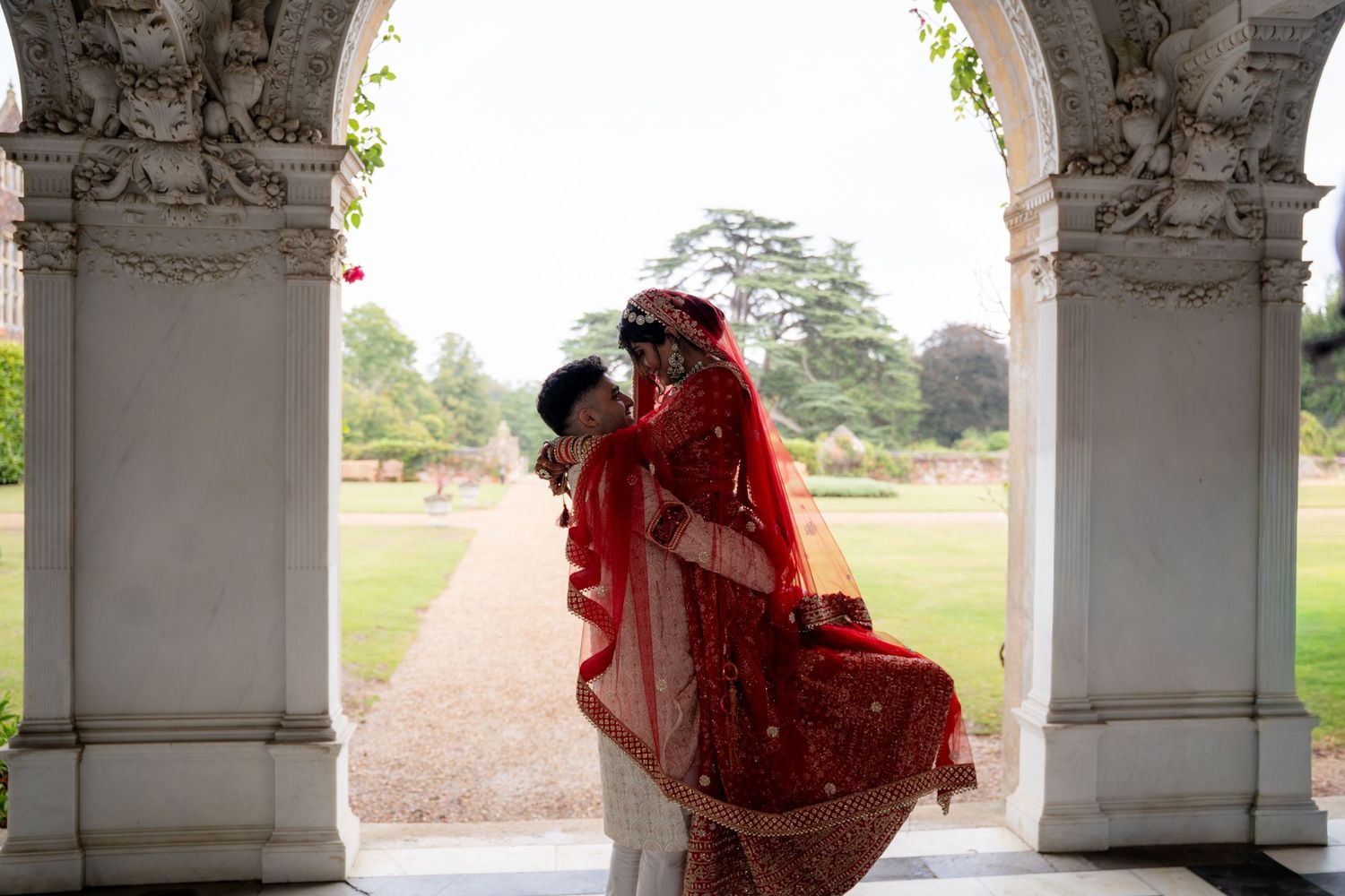 Couple in traditional red Indian wedding attire embracing under a white stone arch in a lush garden.