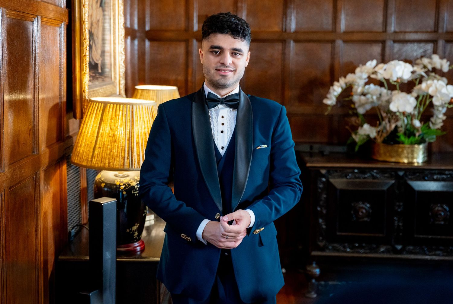Groom in navy blue tuxedo with bow tie posing in elegant wood-paneled room with chandelier.