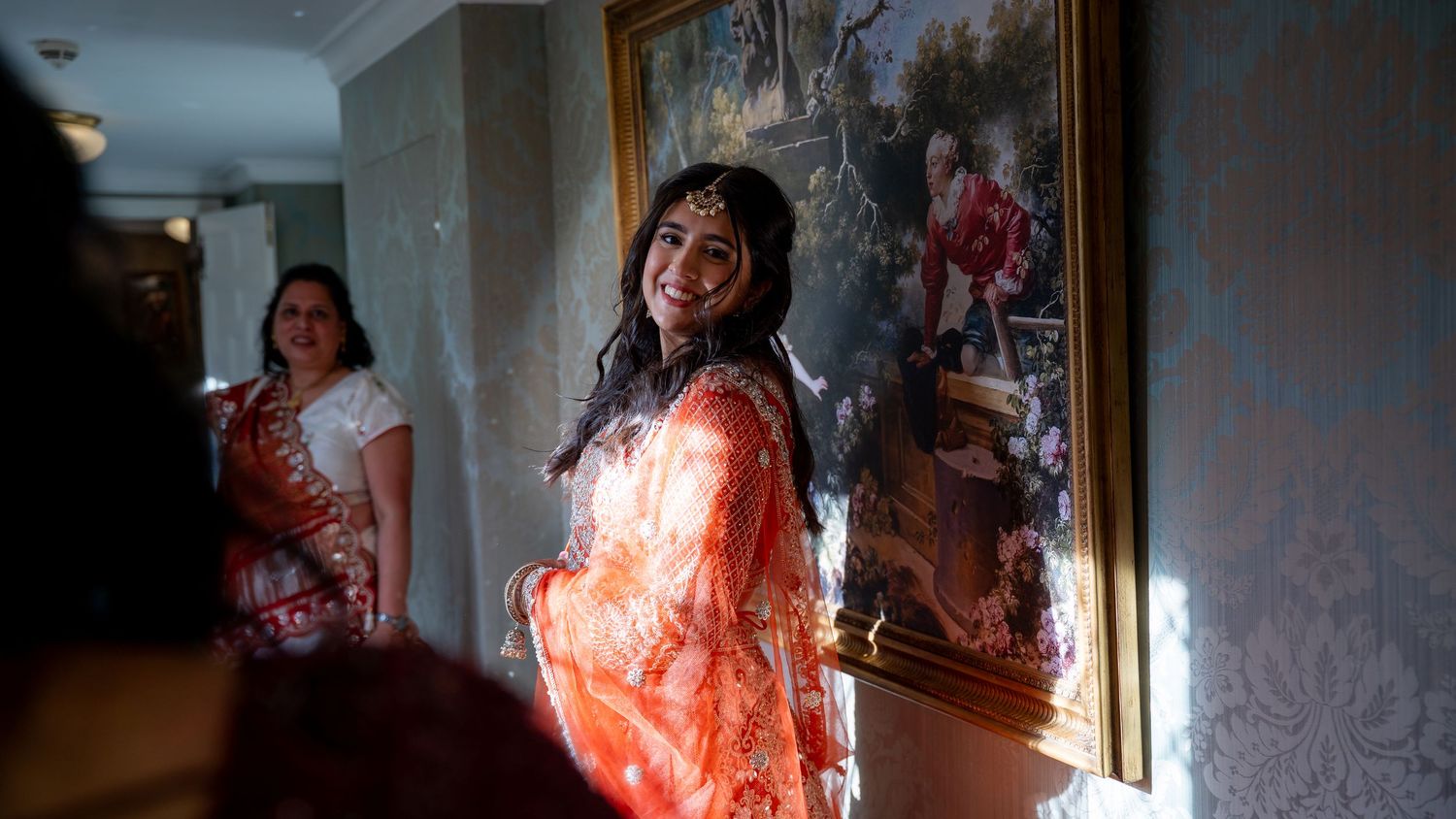A smiling bride in a vibrant orange traditional outfit poses indoors near a decorative painting during wedding preparations.